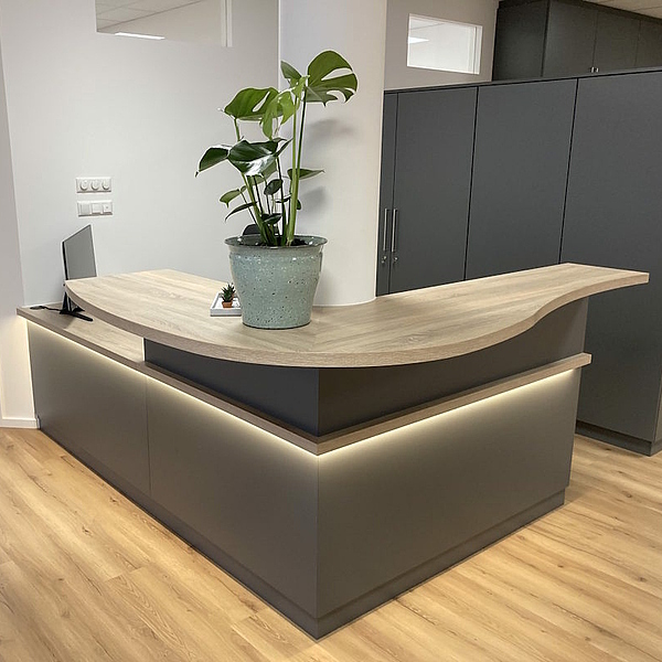 Praxiseinrichtung nach Maß für eine Physiopraxis: Empfangstheke in Anthrazit mit indirekter Beleuchtung, gefertigt von Hofmann Möbel – Ladenbau und Praxismöbel vom Hersteller.